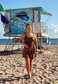 Jeune femme marchant pieds nus sur une plage de sable près d'une tour de sauveteur, avec l'océan et un ciel bleu en arrière-plan.