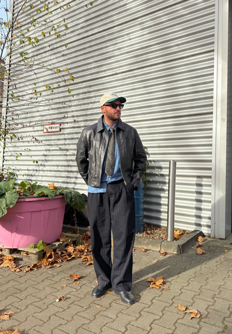 Schwarze Lederjacke, Jeanshemd, gemusterte Krawatte, Nadelstreifenhose, Baseballkappe und Sonnenbrille, stehend neben einem rosa Blumentopf.