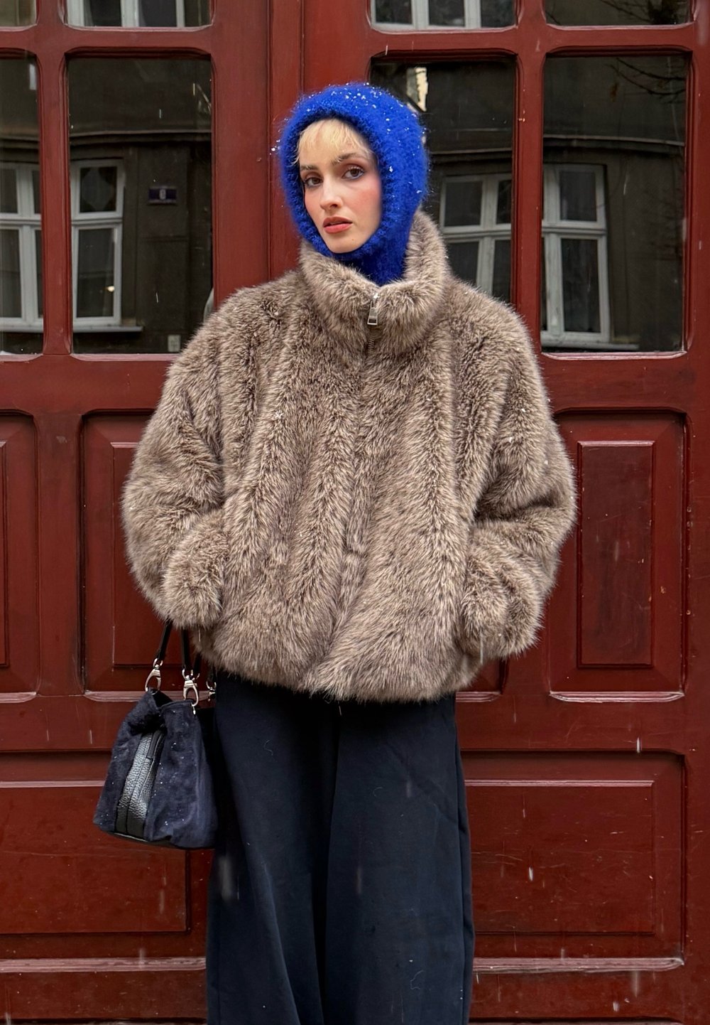 Veste en fausse fourrure marron avec col montant, pantalon large noir, bonnet tricoté bleu et sac foncé avec des accents en cuir devant une porte rouge.