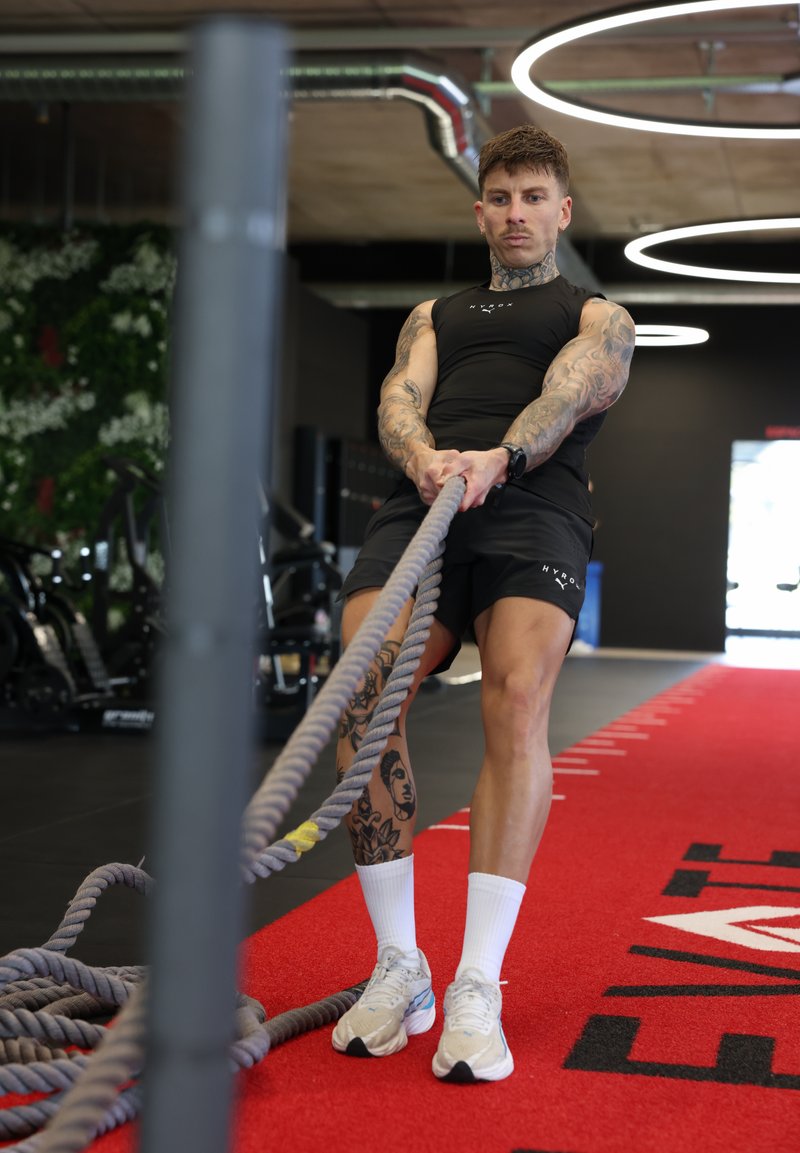 Άνδρας με τατουάζ κάνει άσκηση με battle rope σε κόκκινο γυμναστηριακό δάπεδο, φορώντας μαύρη αθλητική ενδυμασία και λευκά αθλητικά παπούτσια.