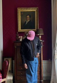 Svart bomberjakke, beige t-skjorte, blå jeans og en rosa caps båret av individ. Mørkerød bakgrunn med et portrett og dekorative elementer.