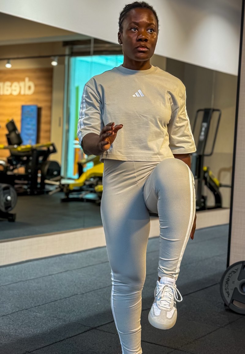 Frau in beigem Adidas-Trainingsoutfit hebt im Fitnessstudio mit Fitnessgeräten und Spiegel im Hintergrund das Knie an.