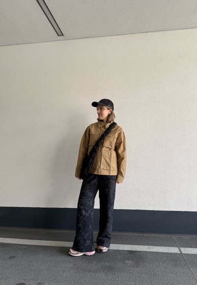 Chaqueta marrón de gran tamaño con dos bolsillos frontales, pantalones anchos negros con estampado, y una gorra negra. El modelo lleva un bolso negro texturizado.