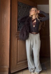 Jeune femme debout dans une porte portant des lunettes, un haut noir zippé sur un soutien-gorge imprimé léopard, un pantalon de survêtement gris et tenant un sac noir en fourrure.