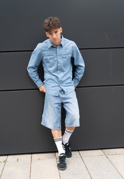 Camisa de denim con cuello abotonado, dos bolsillos en el pecho; pantalones cortos de denim azul claro; zapatillas negras con detalles blancos; calcetines blancos con rayas negras.