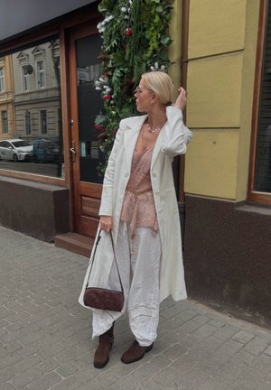 Mujer con abrigo largo blanco y pantalones, blusa rosa de encaje, botas marrones, sosteniendo un bolso burdeos, parada en la acera de la ciudad cerca de una puerta decorada.