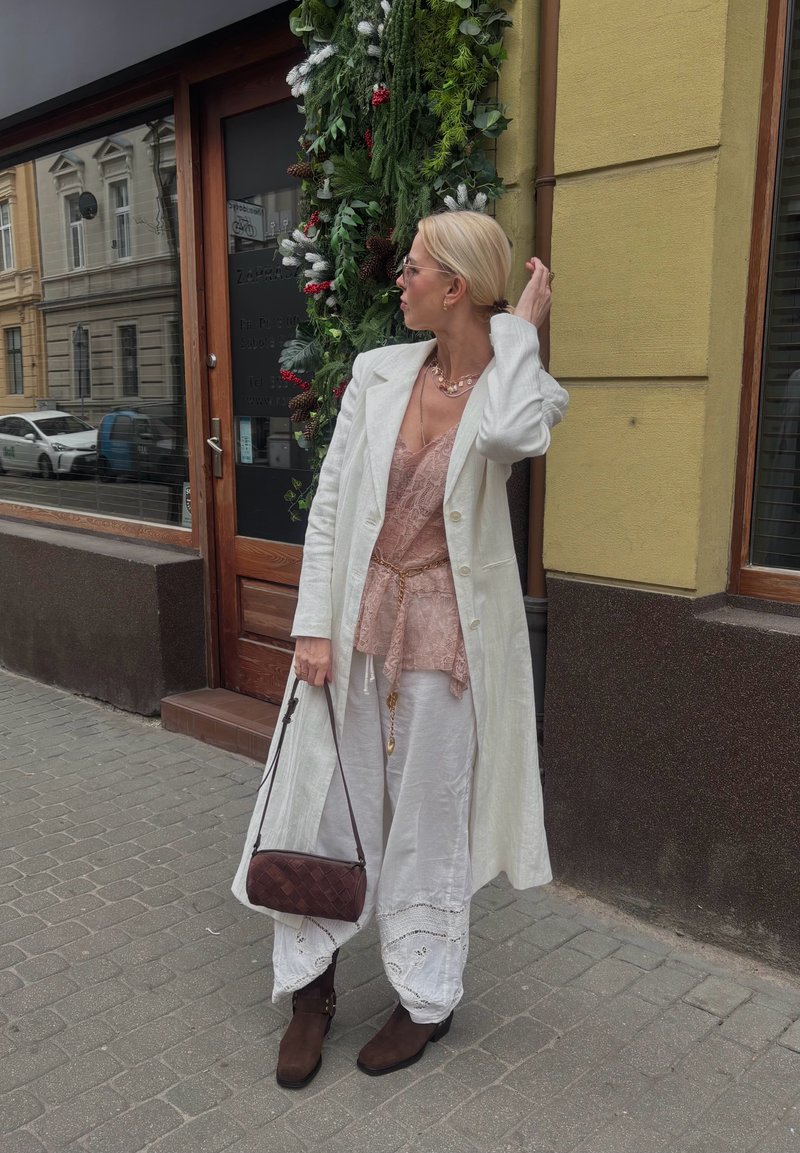 Femeie cu palton lung alb și pantaloni, bluză roz din dantelă, cizme maro, ținând o geantă vișinie, stând pe trotuarul orașului lângă o ușă decorată.