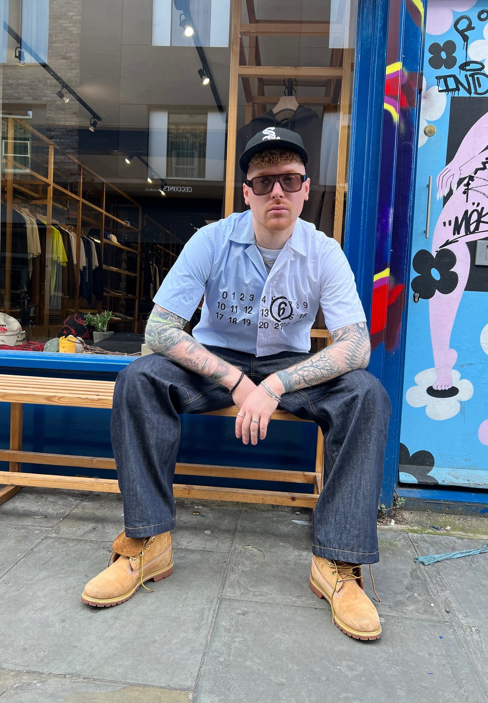 Camisetta azzurro chiaro a maniche corte con stampa grafica nera, jeans baggy blu scuro e stivali da lavoro beige. Seduto su una panchina di legno, all'aperto.