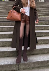 Brauner Mantel, weißer Strickpullover mit Zopfmuster, braune Lederhosen, braune Handtasche und kniehohe braune Strickstiefel auf Steinstufen.