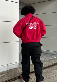Crvena skraćena jakna s natpisom "Alternative Culture STWD" na leđima, uparena s crnim hlačama i bijelim tenisicama s crvenim akcentima.