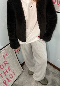 Personne portant une veste noire en peluche, un t-shirt blanc Nike, un pantalon de survêtement gris et des chaussures vertes, debout à côté d'un journal encadré avec un texte rose.