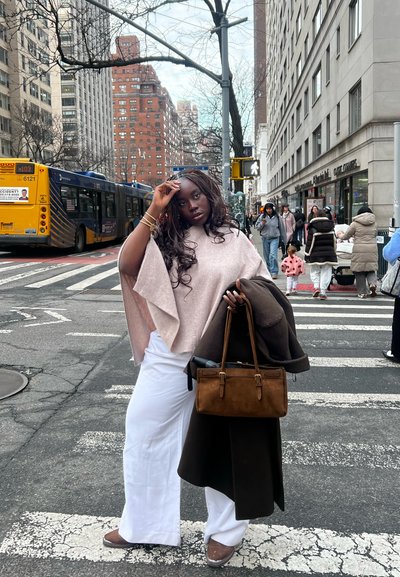 Mujer con poncho rosa pálido y pantalones blancos de pie en un paso de peatones de la ciudad, sosteniendo un abrigo marrón y una bolsa, con un autobús amarillo y edificios urbanos al fondo.