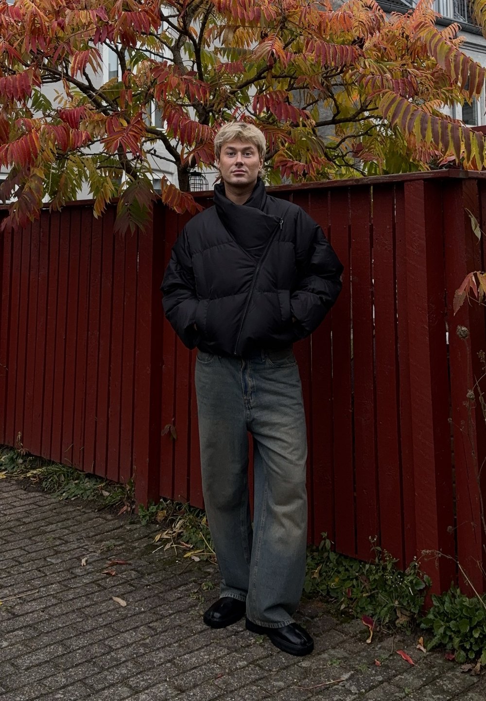 Črno puhovko z diagonalnim zadrgo, ohlapne svetlo modre kavbojke in črne čevlje. Rdeča lesena ograja in jesenske liste v ozadju.