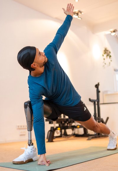 Atleta con una camiseta de manga larga azul y pantalones cortos negros realiza una postura de yoga sobre una esterilla, utilizando una pierna prostética metálica y zapatillas blancas.
