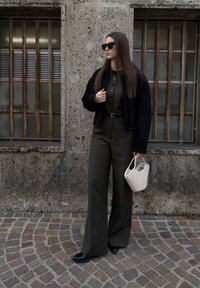 Femme portant des lunettes de soleil foncées, une veste noire, une combinaison olive, des chaussures noires, tenant un sac à main blanc, debout sur une rue pavée avec un mur en pierre en arrière-plan.