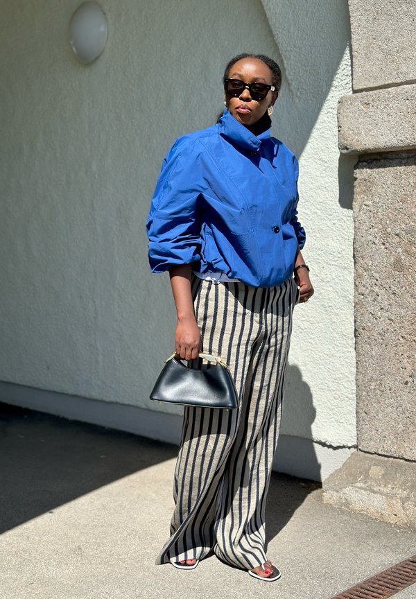 Donna con giacca blu, occhiali da sole neri, pantaloni a gamba larga a righe, tiene una borsa nera, in piedi alla luce del sole vicino a un muro chiaro.