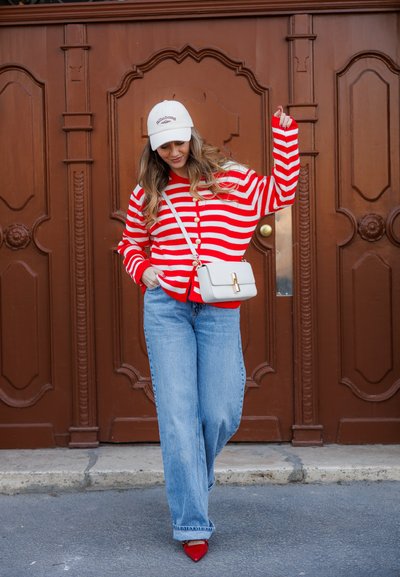 Mujer con suéter a rayas rojas y blancas, jeans azules, zapatos rojos, gorra blanca y bolso blanco cruzado, de pie con un brazo levantado junto a una puerta marrón.