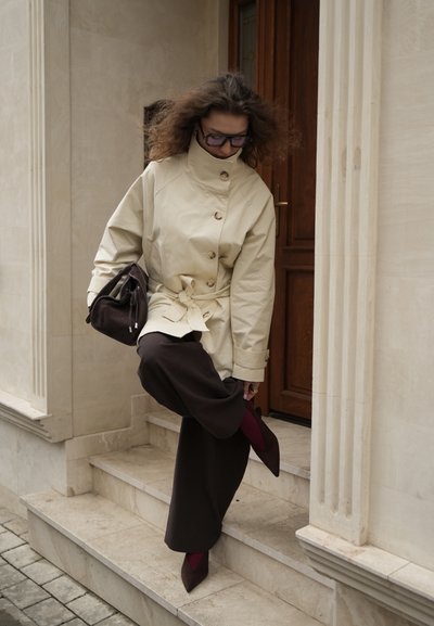 Mujer con chaqueta beige y pantalones anchos oscuros ajustándose un zapato puntiagudo burdeos mientras está de pie en unas escaleras de piedra al aire libre.