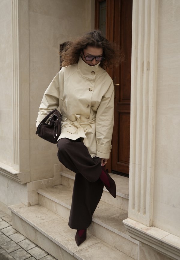 Žena v béžovej bunde a tmavých širokých nohaviciach si upravuje bordó špicatú topánku, stojac na vonkajších kamenných schodoch.