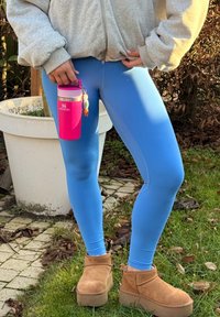 Personne portant un sweat à capuche gris, des leggings bleus et des bottes compensées couleur beige debout sur l'herbe, tenant un thermos Stanley rose avec un porte-clés.