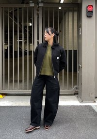 Černá bunda na zip přes olivově zelený svetr, kombinovaná s širokými černými kalhotami a hnědými botami, stojící u kovové brány.