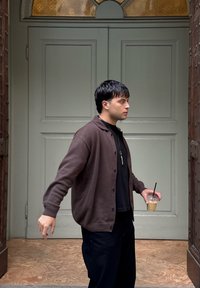 Hnědý cardigan přes černé tričko, stojící před zelenými dveřmi, držící iced coffee. Viditelné texturované dřevěné akcenty.