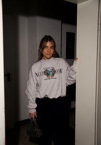 Siva dukserica s natpisom "Yellowstone" i grafikom, crne hlače, drži malu crnu torbicu, stoji na otvorenim vratima.