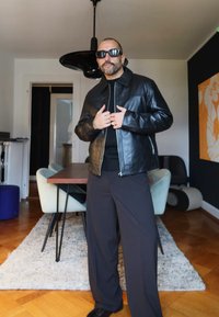 Svart skinnjakke med høy krage, brukt over en svart genser. Vide mørke bukser og svarte sko. Moderne interiør i bakgrunnen.