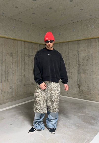 Sudadera negra de gran tamaño, pantalones cortos de leopardo y vaqueros azul holgados. Un gorro rojo y gafas de sol completan el look. Fondo de hormigón.