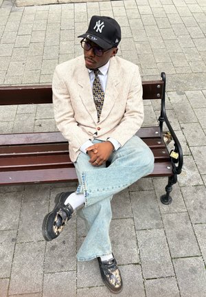 Beige, textured blazer over a white shirt, patterned tie, light blue jeans, black shoes with a distressed look, and a black cap with a logo.