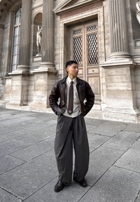 Bruine leren jas, beige overhemd, donkerbruine stropdas, oversized grijze broek en zwarte schoenen tegen een achtergrond van stenen zuilen en sculpturen.