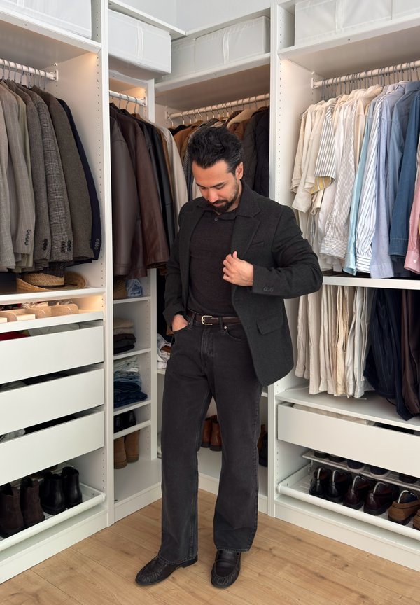 Homme portant des vêtements foncés debout dans un dressing avec des vestes, chemises, chaussures et tiroirs organisés sur des étagères blanches.