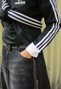 Juodas Adidas treniruoklių švarkas su baltais juostomis, derinamas su tamsiomis džinsinėmis kelnėmis. Sijonų apvadai balti, rodo sklandų tekstūrą ir ženklinimo detalę.