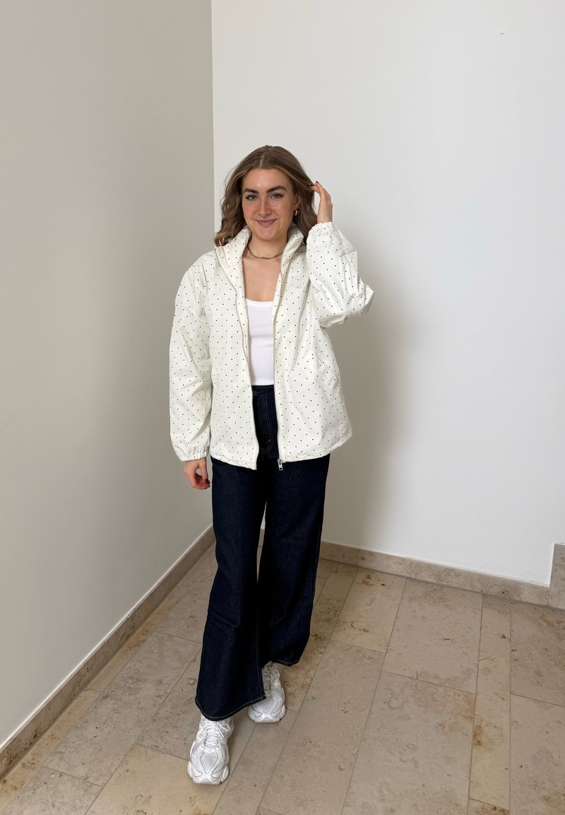 Donna in piedi all'interno, indossa una giacca bianca a pois, una maglietta bianca, jeans scuri a gamba larga e sneakers bianche, sorride con una mano che le tocca i capelli.