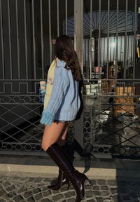 Femme en pull bleu, écharpe jaune et bottes noires montant jusqu'aux genoux, tenant une tasse de café, marchant devant une grille en métal sur des pavés sous le soleil.