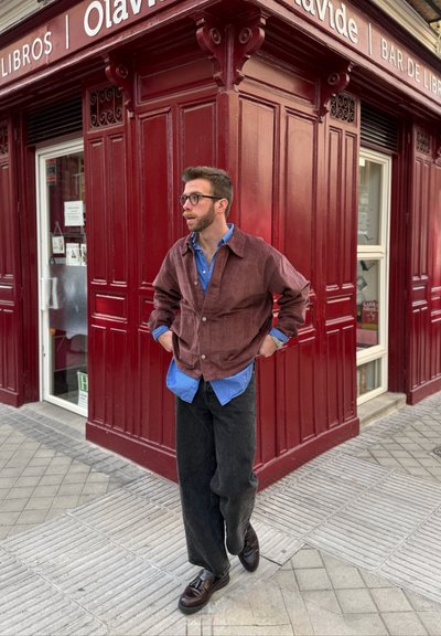 Hombre con gafas, chaqueta marrón, camisa azul y pantalones negros, de pie con las manos en los bolsillos frente a la esquina de una librería roja.