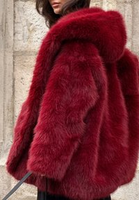 Rød kunstpelsjakke med hætte, der har et massivt design, tyk tekstur og blød finish. Lange ærmer og løs pasform, båret over et sort outfit.