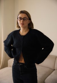 Melnā pūkaina kardigāna ar apaļu kakla izgriezumu un priekšējiem pogām, kombinēts ar augsto jostasvietu tumšajiem džinsiem, mīksta tekstūra un brīvs griezums.