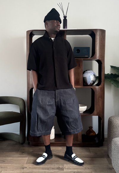 Hombre con gorro negro, camiseta negra de manga corta y pantalones cortos vaqueros holgados, de pie con las manos en los bolsillos en una habitación moderna.