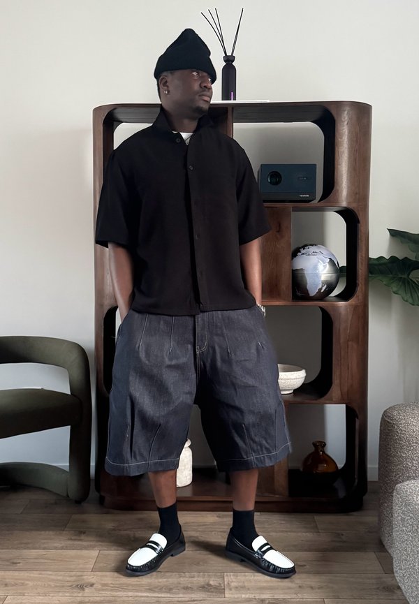 Muž v čiernej čiapke, čiernej krátkorerukej košeli a voľných rifľových šortkách stojí s rukami v náprsných vreckách v moderne vybavenej miestnosti.