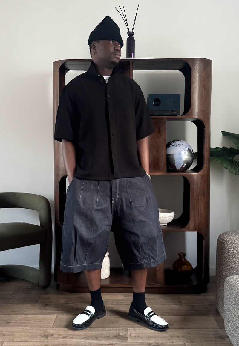 Homme portant un bonnet noir, une chemise noire à manches courtes et un short en jean ample, debout les mains dans les poches dans une pièce moderne.