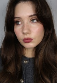 Närbild av en person med långt, mörkbrunt hår, ljus hy, fräknar och en guldsnäppa. Klädd i en grå knappad tröja och ett diskret läppstift.