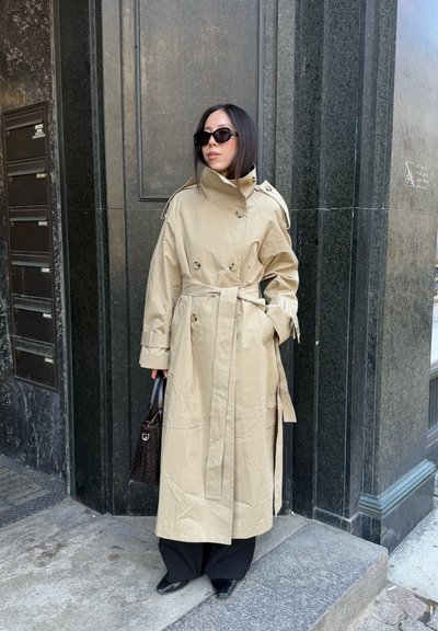 Mujer con gabardina beige, gafas de sol negras, sosteniendo un bolso oscuro, de pie junto a la esquina de un edificio de mármol negro.