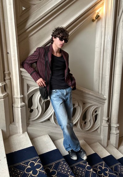 Joven con gafas de sol, chaqueta a rayas, camiseta negra y jeans apoyado en una barandilla de piedra ornamentada en unas escaleras con moqueta.