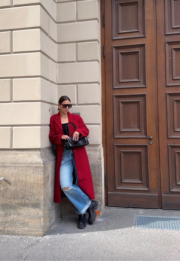 Femme en manteau rouge, jean déchiré et bottes noires appuyée contre un coin de bâtiment, tenant un sac à main noir près de grandes portes en bois.