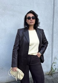 Brown leather blazer over a light sweater, paired with dark pants. Holding a cream-colored, textured handbag. Rounded sunglasses complete the look.