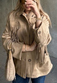 Oseba nosi bež suknjič s frfotom iz semiša, drži usklajen bež ročnjak, na zapestju ima zlato uro in zapestnico.