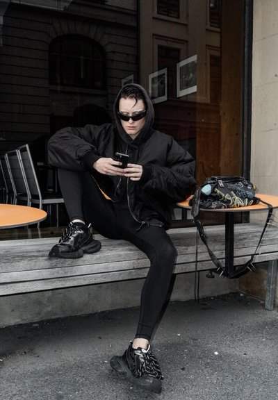 Chaqueta bomber negra, leggings negros y zapatillas gruesas. Sosteniendo un smartphone, sentado en un banco de madera al lado de una mesa naranja.