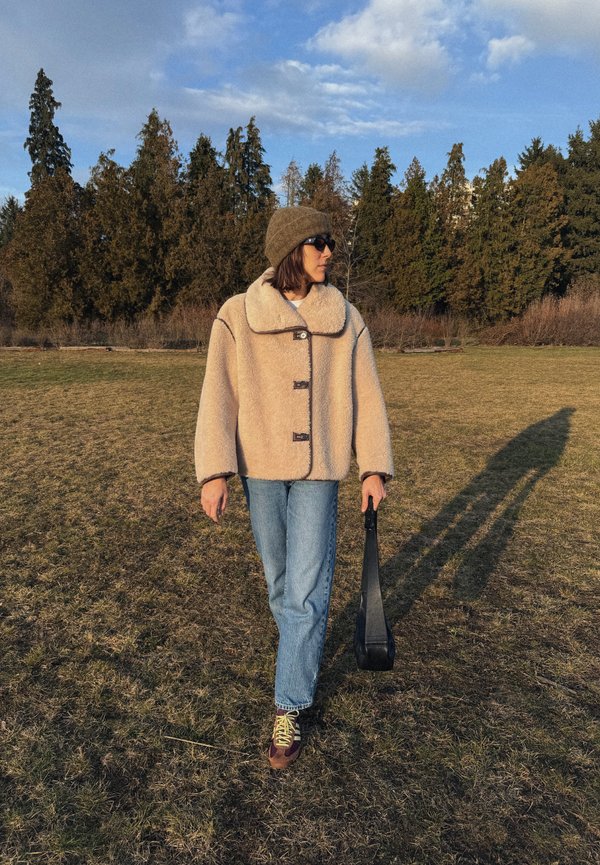 Mujer con abrigo beige, gorro marrón, gafas de sol y vaqueros caminando sobre la hierba en un parque, sosteniendo un bolso negro en su mano derecha.