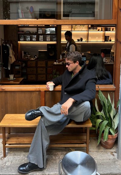 Hombre con gafas de sol y abrigo negro se sienta en un banco de madera fuera de una cafetería, sosteniendo una taza de café, con una planta y un barista visibles en el interior.